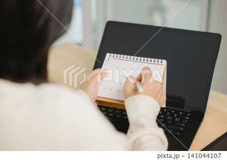 Hand using pen on paper calendar at office desk to schedule date, woman planning workflow with laptop computer, organizing monthly agenda and managing busy business project timeline 141044107
