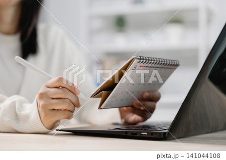 Hand with pen on calendar paper at office desk planning schedule date, while woman holds monthly organizer near open laptop computer to manage important work appointments 141044108
