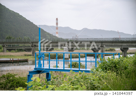 雨が降る曇り空の下で川沿いに佇む青い水門と遠くに見える高架橋や赤白の煙突の風景 雨が降る曇り空の下で川沿いに佇む青い水門と遠くに見える高架橋や赤白の煙突の風景 141044397