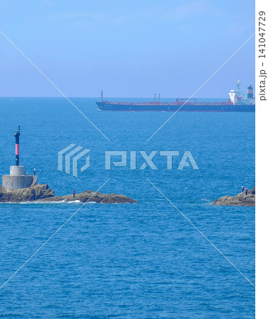 美しい夏の海に浮かぶ岩礁の灯台と磯釣りを楽しむ人々と水平線の貨物船 美しい夏の海に浮かぶ岩礁の灯台と磯釣りを楽しむ人々と水平線の貨物船 141047729
