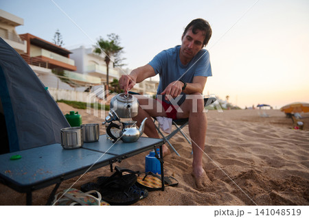 Man making tea while camping on a sandy beach near coastal houses at sunrise 141048519