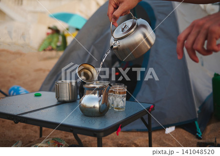 Person pouring tea from a kettle into a teapot at a beach campsite 141048520