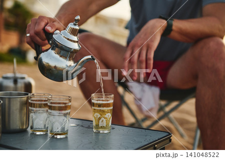Man pouring Moroccan tea outdoors into decorative glasses during relaxed beach gathering Man pouring Moroccan tea outdoors into decorative glasses during relaxed beach gathering 141048522