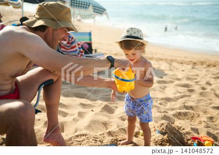 Father and toddler playing with bucket on sunny sandy beach during family summer vacation 141048572