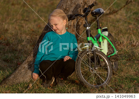 Young blonde girl in a teal shirt and black pants squatting by a tree next to a green bicycle, resting on grass during outdoor play in a park, enjoying a quiet moment Young blonde girl in a teal shirt and black pants squatting by a tree next to a green bicycle, resting on grass during outdoor play in a park, enjoying a quiet moment 141049963