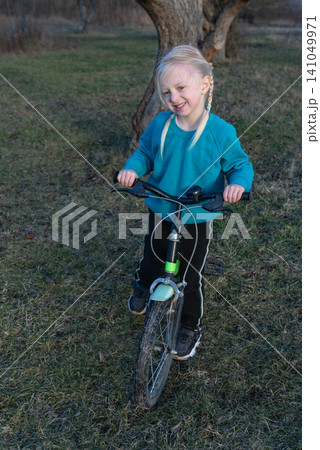 Blond girl in a blue sweatshirt and black pants happily stands with her children's bicycle on green grass, enjoying playful outdoor learning and carefree childhood balance Blond girl in a blue sweatshirt and black pants happily stands with her children's bicycle on green grass, enjoying playful outdoor learning and carefree childhood balance 141049971