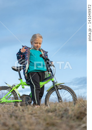 Young girl standing with bright green bicycle on a dry grassy hill, adjusting her jacket and looking down while enjoying a sunny outdoor biking moment in nature Young girl standing with bright green bicycle on a dry grassy hill, adjusting her jacket and looking down while enjoying a sunny outdoor biking moment in nature 141049972