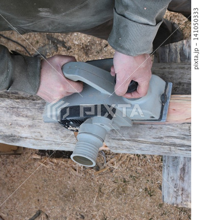 A pair of male hands confidently guides a grey electric planer down the length of an old log, removing the top layer of wood; high-angle view. Fresh sawdust and wood shavings lie scattered around. 141050333
