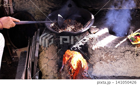 Hand stirring coffee beans in black pan over rustic clay stove with glowing fire, rising smoke and warm orange light during traditional roasting process indoors Hand stirring coffee beans in black pan over rustic clay stove with glowing fire, rising smoke and warm orange light during traditional roasting process indoors 141050426