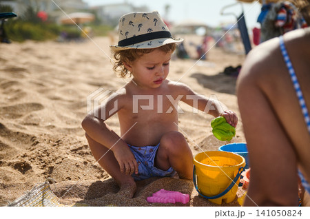 Toddler playing with bucket and toys on sunny beach during family summer vacation Toddler playing with bucket and toys on sunny beach during family summer vacation 141050824