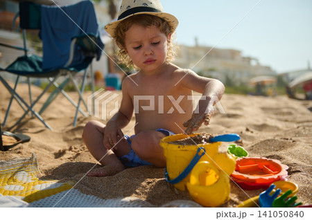 Toddler boy playing with a yellow bucket and sand on a sunny beach during summer vacation Toddler boy playing with a yellow bucket and sand on a sunny beach during summer vacation 141050840