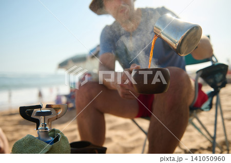 Man pouring hot coffee into wooden cup at the beach during a relaxed camping morning Man pouring hot coffee into wooden cup at the beach during a relaxed camping morning 141050960