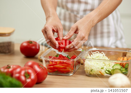 Woman's hands placing chopped fresh tomatoes into a glass food container, preparing healthy meal preps on a wooden kitchen counter Woman's hands placing chopped fresh tomatoes into a glass food container, preparing healthy meal preps on a wooden kitchen counter 141051507