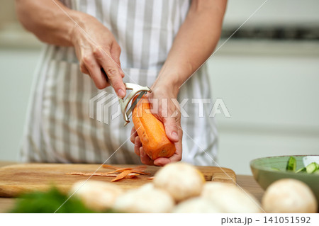 Hands using a peeler to prepare a fresh carrot on a wooden cutting board, making a healthy meal at home Hands using a peeler to prepare a fresh carrot on a wooden cutting board, making a healthy meal at home 141051592