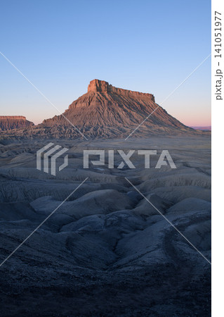 Majestic mountain butte dominates expansive desert landscape utah, revealing ancient rock formation and rugged geology. Its towering peak, striking terrain feature, bathes warm glow of dawn 141051977
