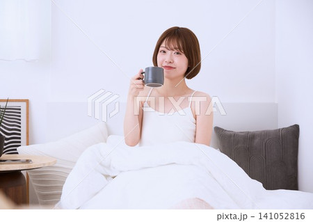 ベッドの上でマグカップを持ちくつろぐ若い女性 ベッドの上でマグカップを持ちくつろぐ若い女性 141052816