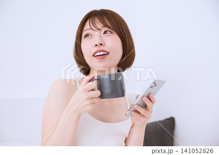 スマートフォンとマグカップを持ちくつろぐ笑顔の若い女性 スマートフォンとマグカップを持ちくつろぐ笑顔の若い女性 141052826
