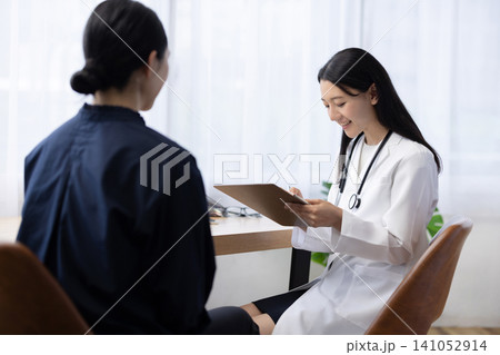 患者の診察中にカルテに記入する若い日本人の女性医師 患者の診察中にカルテに記入する若い日本人の女性医師 141052914