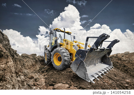 A universal excavator loader is working at a construction site. The loader is digging into dirt while clouds fill the sky above A universal excavator loader is working at a construction site. The loader is digging into dirt while clouds fill the sky above 141054258