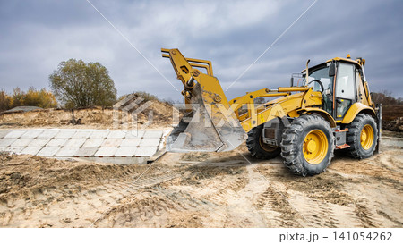 A universal excavator loader operates on a construction site. It moves materials and digs into sandy soil under a cloudy sky at an outdoor location A universal excavator loader operates on a construction site. It moves materials and digs into sandy soil under a cloudy sky at an outdoor location 141054262
