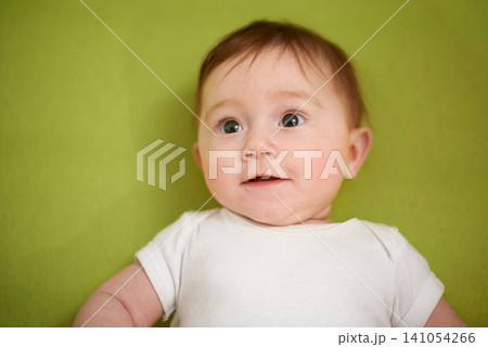 Happy, cute and closeup of baby in studio playing for child development and curious face. Smile, sweet and zoom of young girl kid, infant or newborn relaxing and laying isolated by green background. 141054266