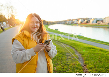 woman texting on phone in relaxed outdoor park near river with cotton and plastic case, casual and serene, yellow and green tones, frontal view, lifestyle and technology context. woman texting on phone in relaxed outdoor park near river with cotton and plastic case, casual and serene, yellow and green tones, frontal view, lifestyle and technology context. 141055035