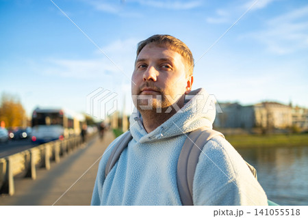 man looking up in urban riverside setting with thoughtful expression and backpack straps visible on concrete road near metal railing, exploring city life and travel lifestyle. 141055518