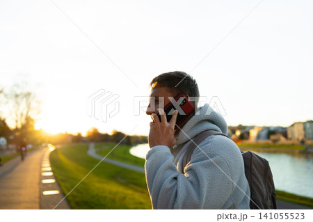 man talking on phone in relaxed park setting at sunset side view with backpack and clothing in natural light and gray and red tones, for editorial and educational visual. 141055523
