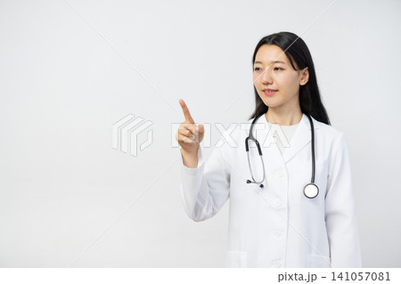 白い背景の前で空中を指さして微笑む若い日本人の女性医師 白い背景の前で空中を指さして微笑む若い日本人の女性医師 141057081