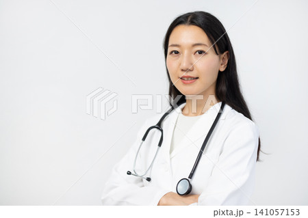 白い背景の前で腕組みをしてカメラ目線で微笑む白衣を着た若い日本人の女性医師 141057153