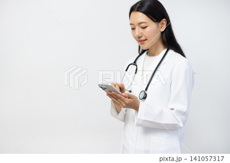 白い背景の前でスマートフォンを操作しながら微笑む白衣を着た若い日本人の女性医師 141057317
