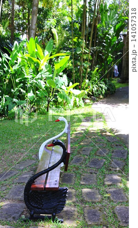 Ornamental swan-shaped bench beside stone garden path surrounded by lush tropical foliage, sunlit lawn, palm trunks, broadleaf plants, serene resort-style outdoor seating Ornamental swan-shaped bench beside stone garden path surrounded by lush tropical foliage, sunlit lawn, palm trunks, broadleaf plants, serene resort-style outdoor seating 141057318