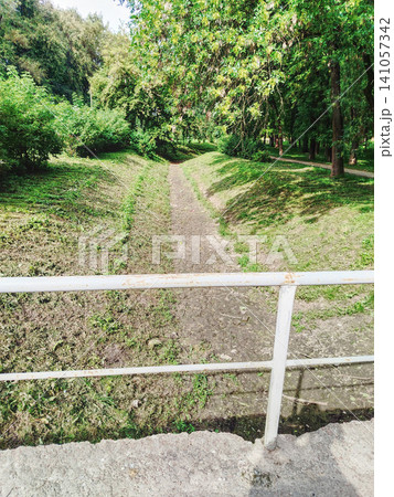 Sunny Green Park Pathway Through Trees With White Metal Railing Over Small Dry Streambed. 141057342