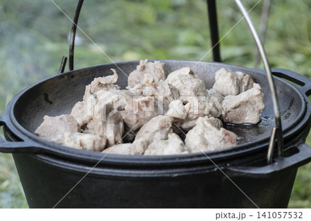 Cooking delicious meat in a large cast-iron cauldron over an open campfire in nature. A rustic outdoor meal preparation scene, showcasing traditional cooking methods during a hike or camping Cooking delicious meat in a large cast-iron cauldron over an open campfire in nature. A rustic outdoor meal preparation scene, showcasing traditional cooking methods during a hike or camping 141057532