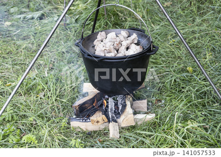 Cooking delicious meat in a large cast-iron cauldron over an open campfire in nature. A rustic outdoor meal preparation scene, showcasing traditional cooking methods during a hike or camping Cooking delicious meat in a large cast-iron cauldron over an open campfire in nature. A rustic outdoor meal preparation scene, showcasing traditional cooking methods during a hike or camping 141057533