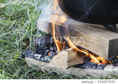 Close-up view of a small campfire made of stacked wood logs and charcoal on green grass, actively burning with visible flames and rising smoke, heating the bottom of a black pot for cooking. 141057545