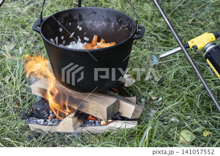 Black cast iron pot with carrots and onions, hanging over a small campfire built on the green grass. Perfect for rustic outdoor cooking and camping. Black cast iron pot with carrots and onions, hanging over a small campfire built on the green grass. Perfect for rustic outdoor cooking and camping. 141057552