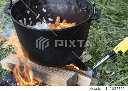 Black cast iron pot with carrots and onions, hanging over a small campfire built on the green grass. Perfect for rustic outdoor cooking and camping. Black cast iron pot with carrots and onions, hanging over a small campfire built on the green grass. Perfect for rustic outdoor cooking and camping. 141057553