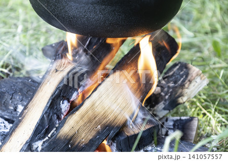 Close-up view of a small campfire made of stacked wood logs and charcoal on green grass, actively burning with visible flames and rising smoke, heating the bottom of a black pot for cooking. 141057557