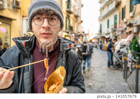Naples, italy. December 27, 2024. Young man holding a paper cone with fried seafood using chopsticks while walking along the famous spaccanapoli street in naples Naples, italy. December 27, 2024. Young man holding a paper cone with fried seafood using chopsticks while walking along the famous spaccanapoli street in naples 141057679