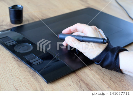 Designer's hand, wearing a black two-fingered art glove, draws with a digital stylus on a black graphics tablet. Digital illustration process 141057711