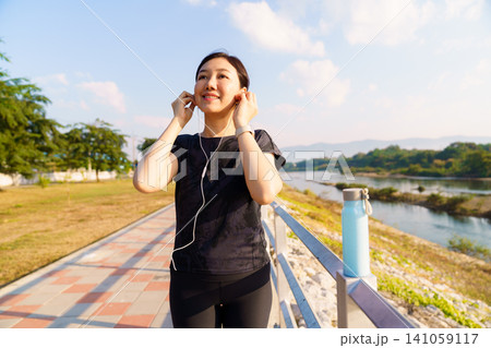 Woman wearing earphones preparing for outdoor exercise near river. Fitness and healthy lifestyle concept with workout, wellness and active living. Woman wearing earphones preparing for outdoor exercise near river. Fitness and healthy lifestyle concept with workout, wellness and active living. 141059117