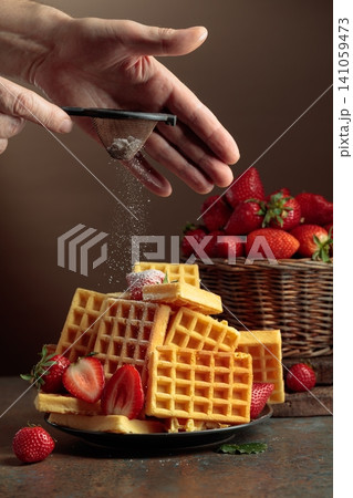 The chef sprinkles powdered sugar on the waffles topped with fresh strawberries. The chef sprinkles powdered sugar on the waffles topped with fresh strawberries. 141059473