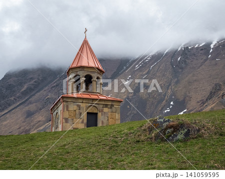 Ancient Stone Bell Tower of Gergeti Trinity Church Against Cloudy Caucasus Mountains in Kazbegi, Georgia. 141059595