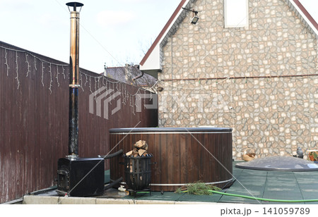Outdoor hot tub with wood exterior and metal stove beside fence, installed on terrace near brick building. Hot tub heated with firewood creating cozy spa atmosphere, backyard relaxation concept, Outdoor hot tub with wood exterior and metal stove beside fence, installed on terrace near brick building. Hot tub heated with firewood creating cozy spa atmosphere, backyard relaxation concept, 141059789