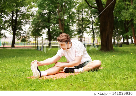 man is stretching on a mat on green grass in the park. This outdoor fitness exercise and daily workout routine is excellent for his overall health. 141059930