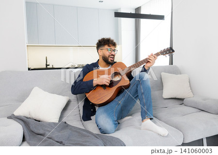 Happy man playing acoustic guitar on sofa 141060003
