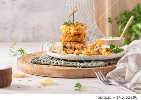A vertical stack of golden crispy cod fritters with lemon and parsley on a rustic wooden board, accompanied by a bowl of creamy dip for a delicious meal A vertical stack of golden crispy cod fritters with lemon and parsley on a rustic wooden board, accompanied by a bowl of creamy dip for a delicious meal 141062180