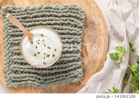 A bowl of creamy white yogurt or sour cream topped with fresh chives sits on a rustic wooden board and a soft green knit cloth with a casual fabric drape 141062186