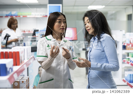 Asian pharmacist assists curious female customer browsing supplements in local retail pharmacy. Professional consultation focuses on product benefits, wellbeing and accurate health information. Asian pharmacist assists curious female customer browsing supplements in local retail pharmacy. Professional consultation focuses on product benefits, wellbeing and accurate health information. 141062389